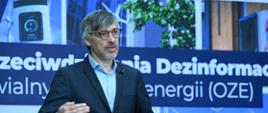 Mężczyzna w garniturze (Krzysztof Bolesta, Sekretarz Stanu w Ministerstwie Klimatu i Środowiska) przemawia do mikrofonu na tle ekranu, na którym wyświetlany jest napis: „Laboratorium przeciwdziałania dezinformacji w obszarze odnawialnych źródeł energii (OZE)”.