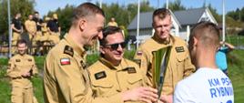 Uroczysta ceremonia wręczenia nagród na świeżym powietrzu. Trzej uśmiechnięci strażacy Państwowej Straży Pożarnej w piaskowych mundurach przekazują duży, złoty puchar zawodnikowi stojącemu tyłem, ubranemu w biało-niebieską koszulkę sportową z numerem 10 i napisem KM PSP Rybnik. W tle na trawniku widać pozostałych uczestników wydarzenia oraz budynki pod pogodnym niebem.