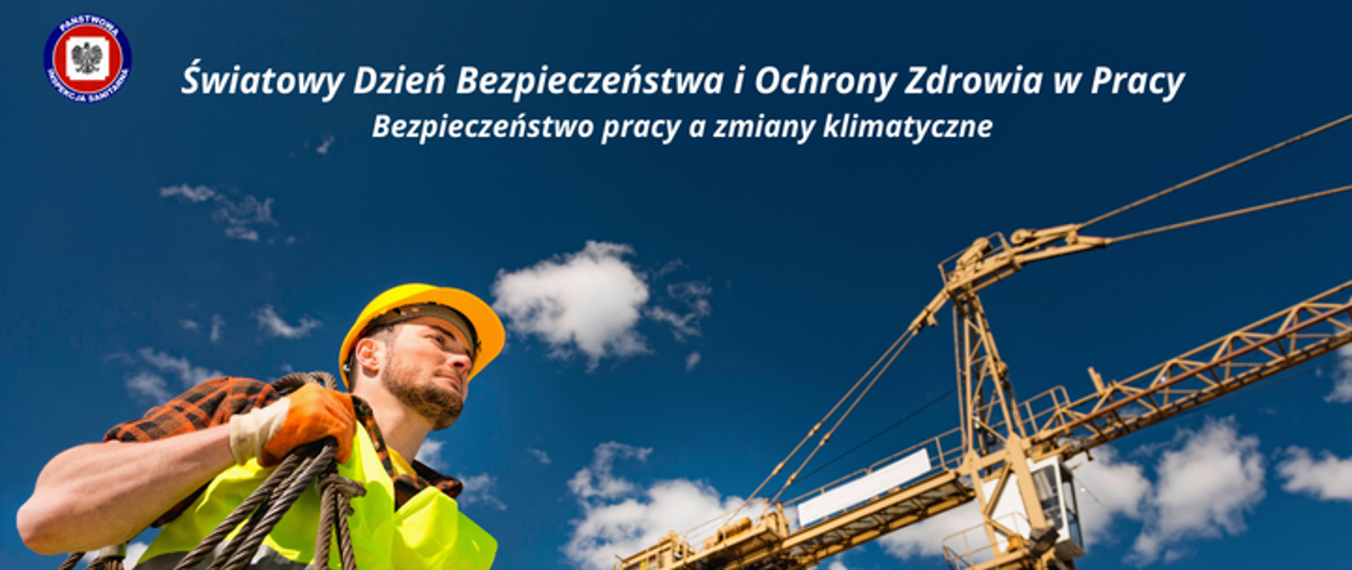 Na tle niebieskiego nieba po prawej żółty żuraw, po lewej Robotnik w stroju roboczym i w kasku z liną na ramieniu. U góry, pośrodku jasny napis Światowy dzień Bezpieczeństwa i Ochrony Zdrowia w Pracy, poniżej Bezpieczeństwo pracy a zmiany klimatyczne. Po jego lewej logo Państwowej Inspekcji Sanitarnej. 