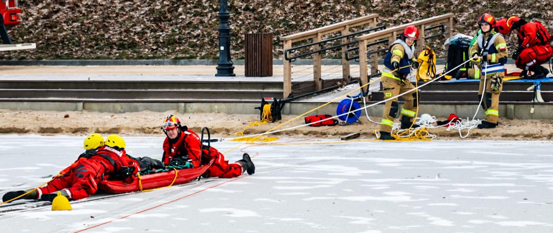 Strażacy wciągają ćwiczących strażaków po lodzie na saniach lodowych.