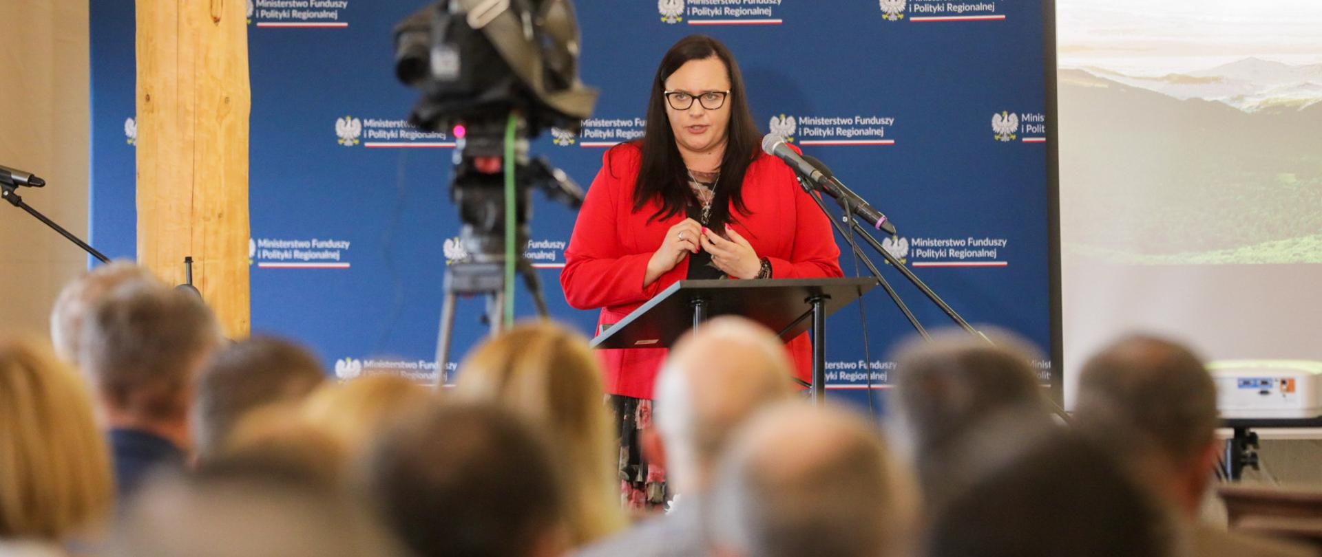 Wiceminister Małgorzata Jarosińska-Jedynak przed mikrofonami na tle ścianki MFiPR. Przed nią publiczność konferencji siedzi na krzesłach.