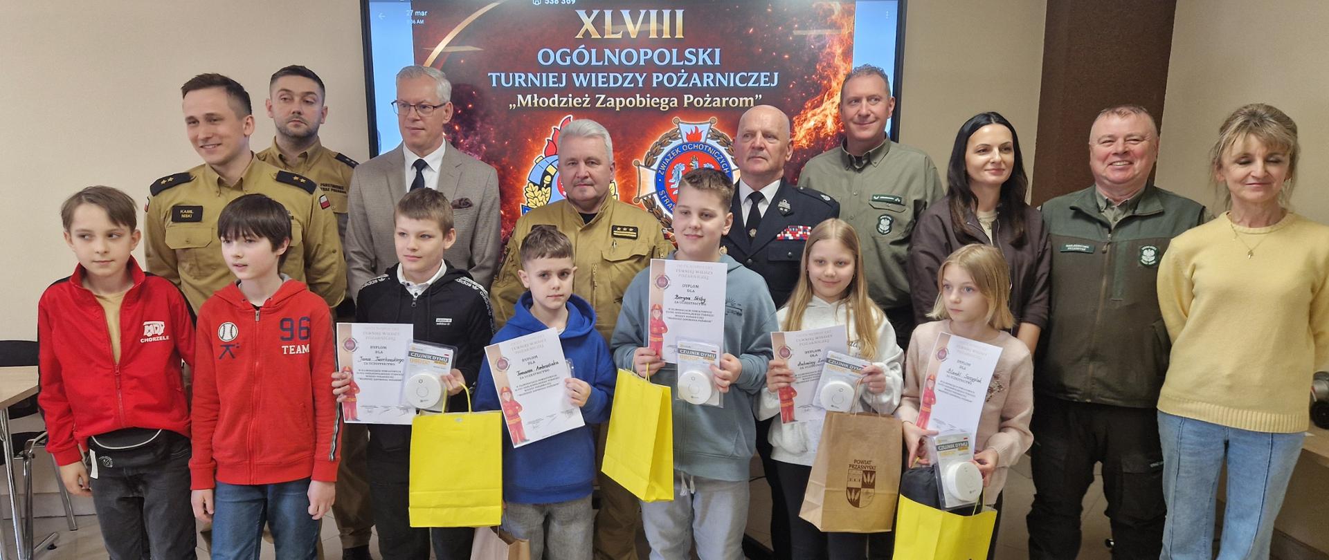 Pamiątkowe zdjęcie uczestników i organizatorów konkursu pożarniczego, wykonane w pomieszczeniu na tle dużego ekranu.
Pierwszy plan: Grupa siedmiorga dzieci w wieku szkolnym stoi w rzędzie. Pięcioro z nich trzyma dyplomy, papierowe torby z upominkami oraz białe, okrągłe czujniki dymu w opakowaniach. Dzieci są ubrane w stroje codzienne – bluzy z kapturem (czerwoną, niebieską, szarą), swetry i spodnie dżinsowe.
Drugi plan: Za dziećmi stoi dziewięć osób dorosłych. Wśród nich znajdują się funkcjonariusze Straży Pożarnej w różnych typach mundurów (piaskowych oraz ciemnym mundurze wyjściowym z odznaczeniami), mężczyźni w zielonych mundurach leśników oraz osoby w strojach cywilnych (garniturze, skórzanej kurtce i żółtym swetrze).
Tło: Centralnym elementem tła jest monitor wyświetlający grafikę z napisem: „XLVIII OGÓLNOPOLSKI TURNIEJ WIEDZY POŻARNICZEJ 'Młodzież Zapobiega Pożarom'”. Na ekranie widoczne są również logotypy straży pożarnej oraz motyw płomieni.
Kompozycja: Wszyscy patrzą w stronę obiektywu i lekko się uśmiechają. Oświetlenie jest jasne, wnętrze wygląda na salę konferencyjną lub świetlicę.