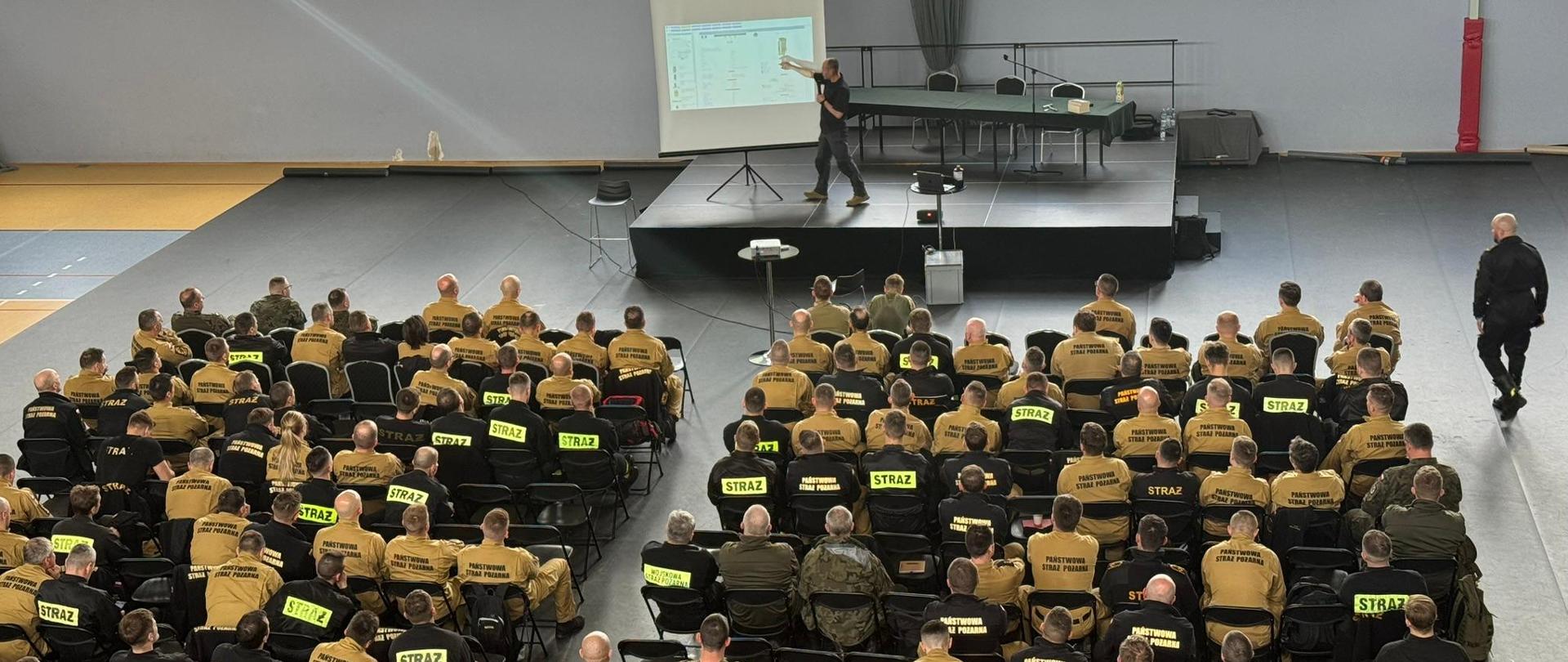 Słuchacze siedzą na krzesłach na sali gimnastycznej w mundurach służbowych. Przed nimi znajduje się podest, na którym znajduje się prowadzący szkolenie mężczyzna. Obok niego stoi rozstawiony ekran, na którym jest wyświetlana prezentacja.