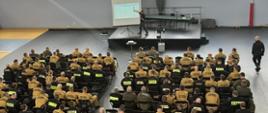 Słuchacze siedzą na krzesłach na sali gimnastycznej w mundurach służbowych. Przed nimi znajduje się podest, na którym znajduje się prowadzący szkolenie mężczyzna. Obok niego stoi rozstawiony ekran, na którym jest wyświetlana prezentacja.