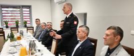 Dowódca w mundurze strażackim przemawia do uczestników spotkania świątecznego siedzących przy wspólnym stole. Przy stole zasiadają zaproszeni goście i funkcjonariusze, a na stole widoczne są świąteczne dekoracje, poczęstunek, kawa i herbata.