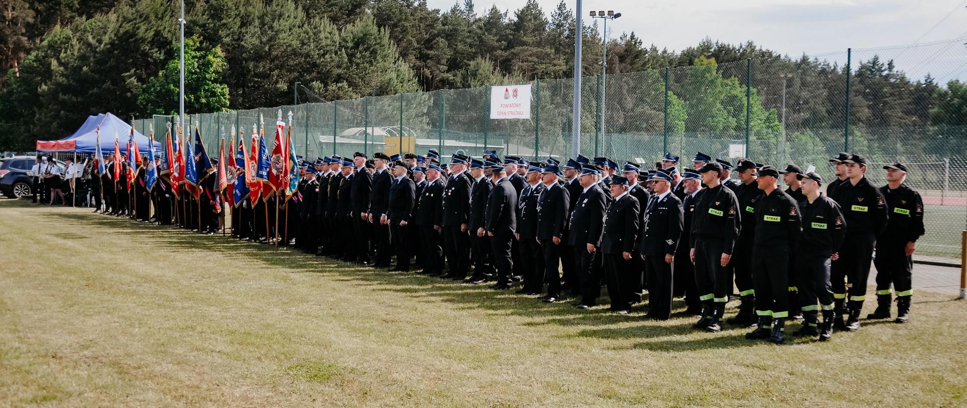 w oddali po lewej orkiestra osp pod namiotem, następnie poczty sztandarowe i pododdziały strażaków i druhów stojących w linii na apelu na trawiastym boisku