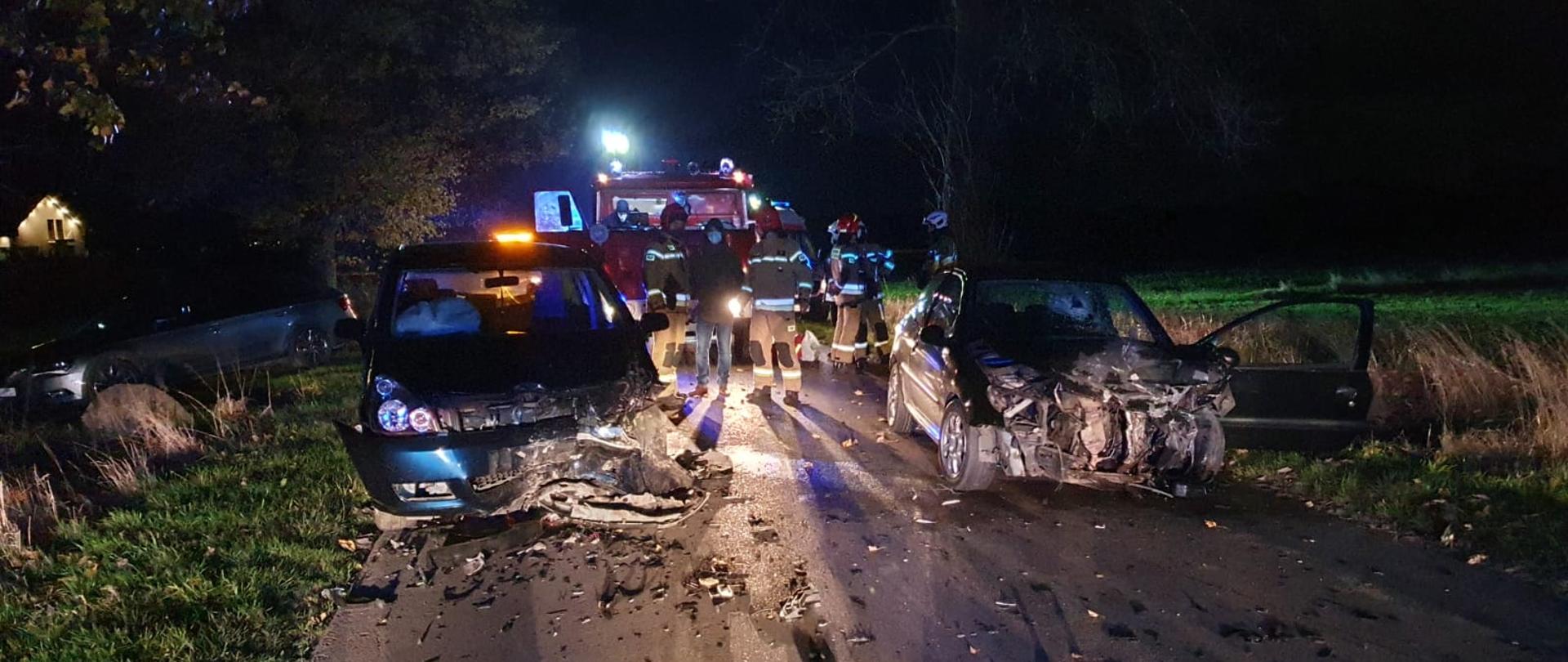 Na zdjęciu w porze wieczornej na jedni znajdują się dwa samochody osobowe po zderzeniu czołowym (rozbite przody) w tle samochody straży pożarnej.