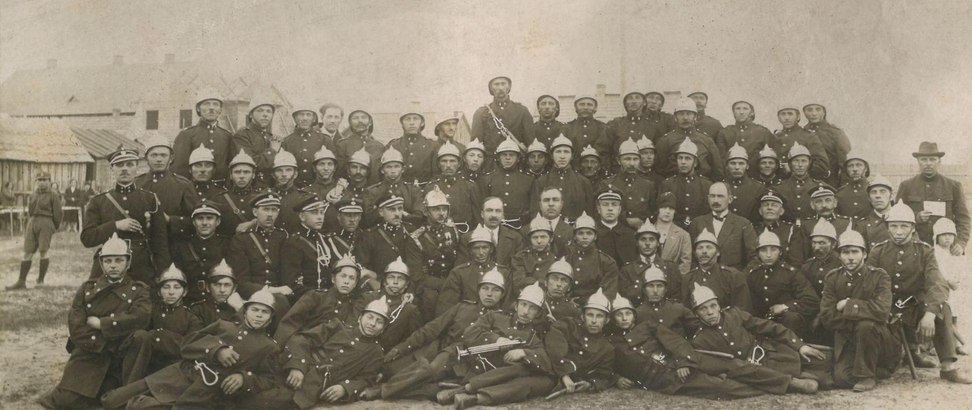 W kilku rzędach ustawionych jest kilkadziesiąt osób. W większości strażacy w galowych mundurach.