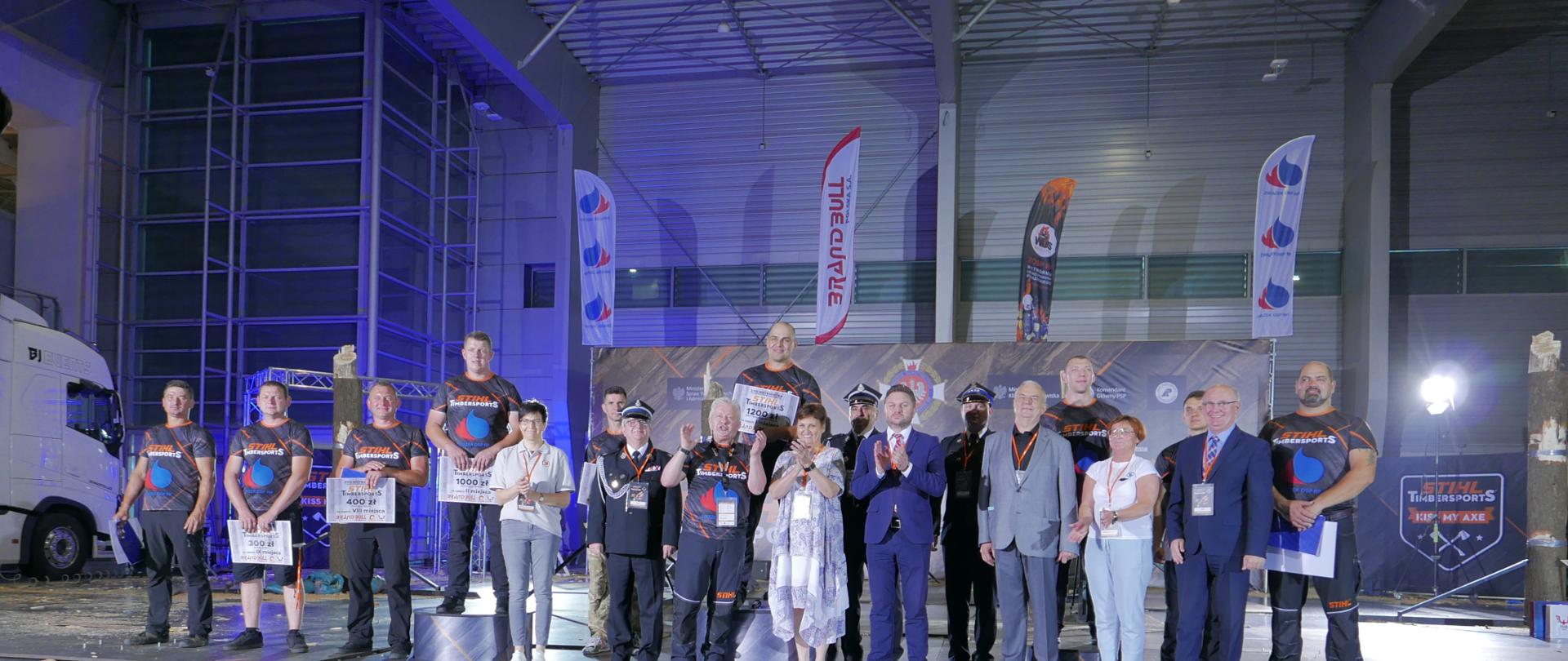 grupa osób stojąca przy podium, wśród nich kobiety oraz strażacy , na podium stoi trzech mężczyzn z pucharami
