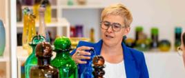 Wiceminister Mrówczyńska w niebieskiej marynarce stoi przy białym stoliku, na którym stoją butelki z kolorowego szkła.