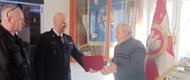 Komendant powiatowy PSP w Żyrardowie wręcza pierwszemu druhowi OSP decyzję o przyznaniu świadczenia ratowniczego. Obok komendanta stoi jego zastępca. W tle wisi sztandar komendy.