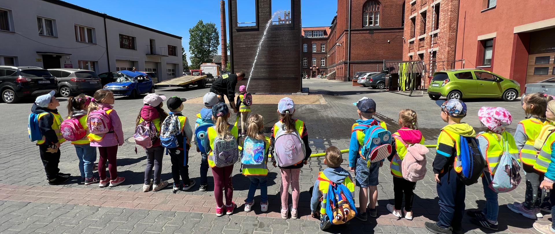 Słoneczny dzień. Niebieskie niebo. Dzieci w żółtych kamizelkach z plecakami na plecach leją wodą z węża strażackiego. W tle budynki komendy. 