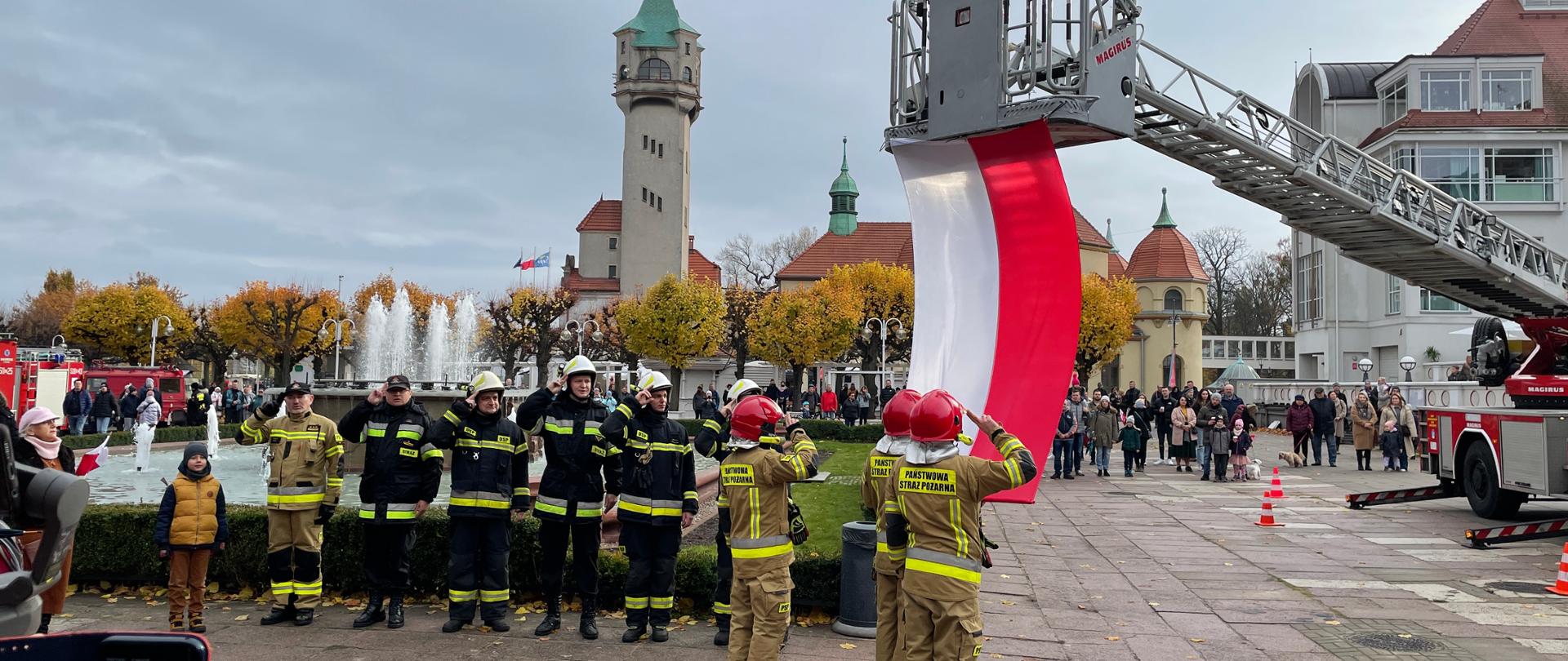 Uroczyste Podniesienie Flagi Państwowej na Placu Zdrojowym. Flaga zaczepiona do kosza drabiny, podtrzymują ją poczet flagowy Obok w szeregu stoją strażacy i oddają honor. W tle widać mieszkańców