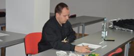 Egzamin recertyfikacyjny. Świetlica Komendy Powiatowej PSP w Rawiczu. Przy stole siedzi członek komisji egzaminacyjnej, który ocenia zadania wykonywane przez egzaminowanych. W ręce trzyma długopis. Przed nim na stole leży sprzęt do udzielania kpp. 