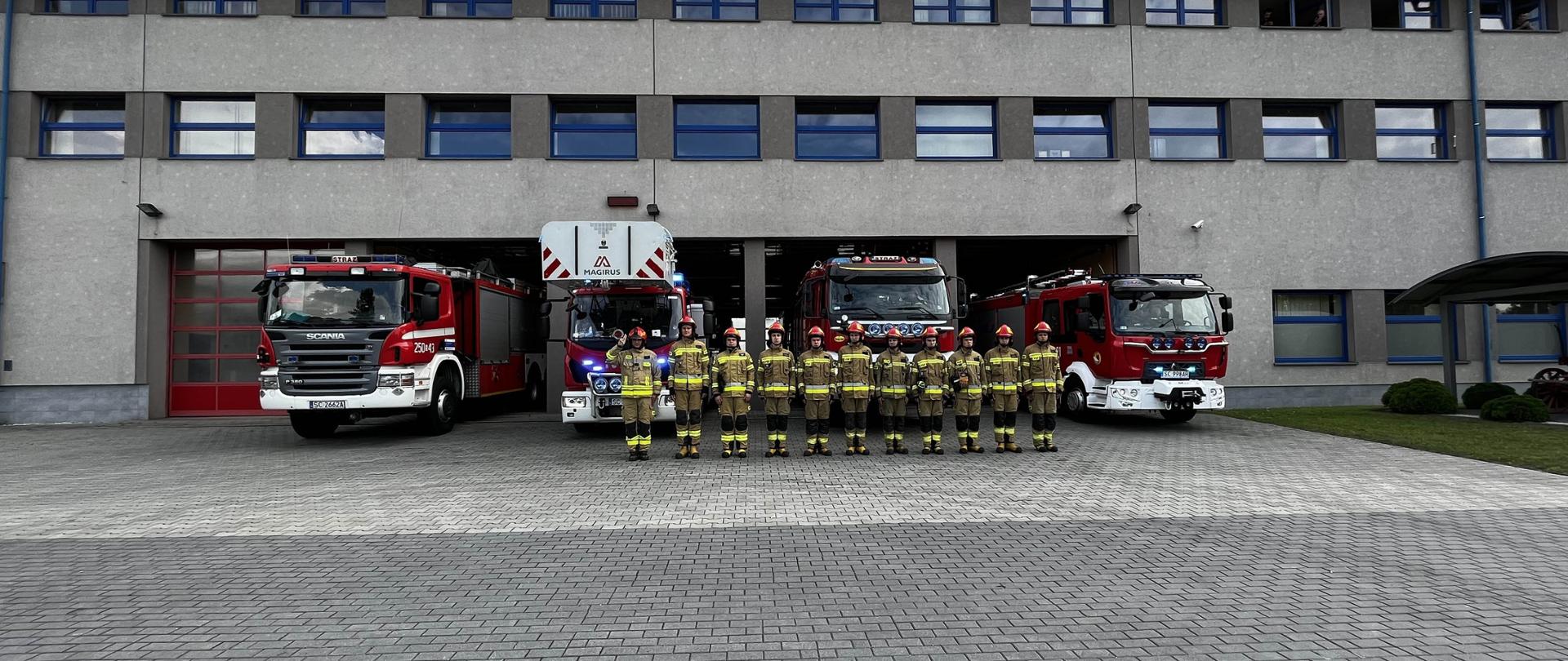 Strażacy w strojach bojowych (11 osób) stoją w szeregu przed garażami Jednostki Ratowniczo-Gaśniczej. Za nimi wystawione wozy bojowe z włączonymi sygnałami świetlnymi