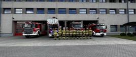 Strażacy w strojach bojowych (11 osób) stoją w szeregu przed garażami Jednostki Ratowniczo-Gaśniczej. Za nimi wystawione wozy bojowe z włączonymi sygnałami świetlnymi