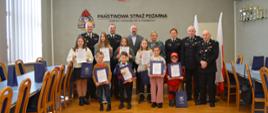 ośmioro dzieci stoi w dwóch rzędach trzymają dyplomu za nimi w jednym rzędzie stoi pięciu strażaków i miedzy nimi jeden pan i jedna pani. zdjęcie wykonane na sali Na ścianie wisi napis Państwowa Straż Pożarna Komenda Powiatowa PSP w Poddębicach