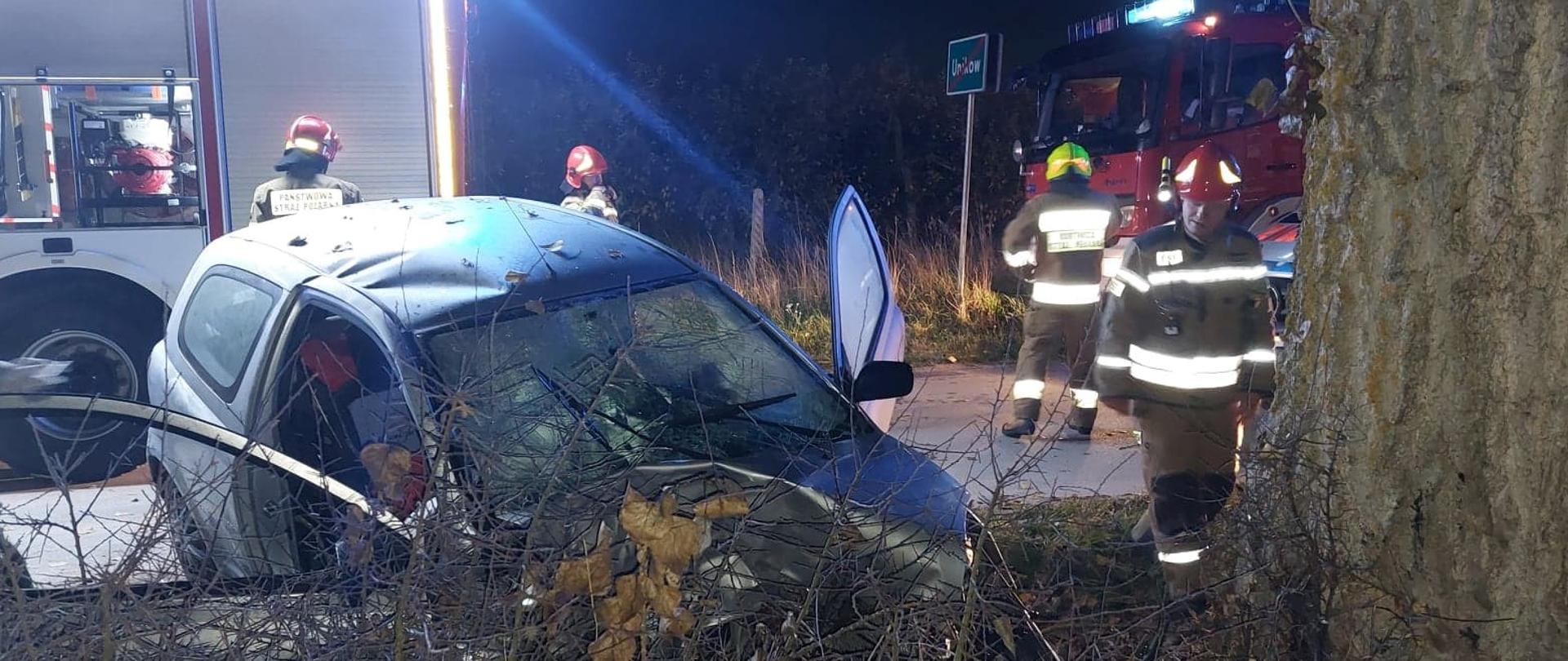 Strażacy prowadzą działania w obrębie wraku pojazdu Toyota Yaris. W tle widać wozy pożarnicze oraz karetkę PRM. 