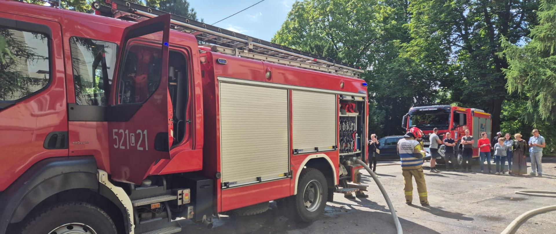 samochody ratowniczo-gaśnicze z rozwiniętą linią wężową, obok stoi kierujący działaniem ratowniczym ubrany w ubraniu specjalnym oraz hełmie.na dalszym planie ewakuowani pracownicy.