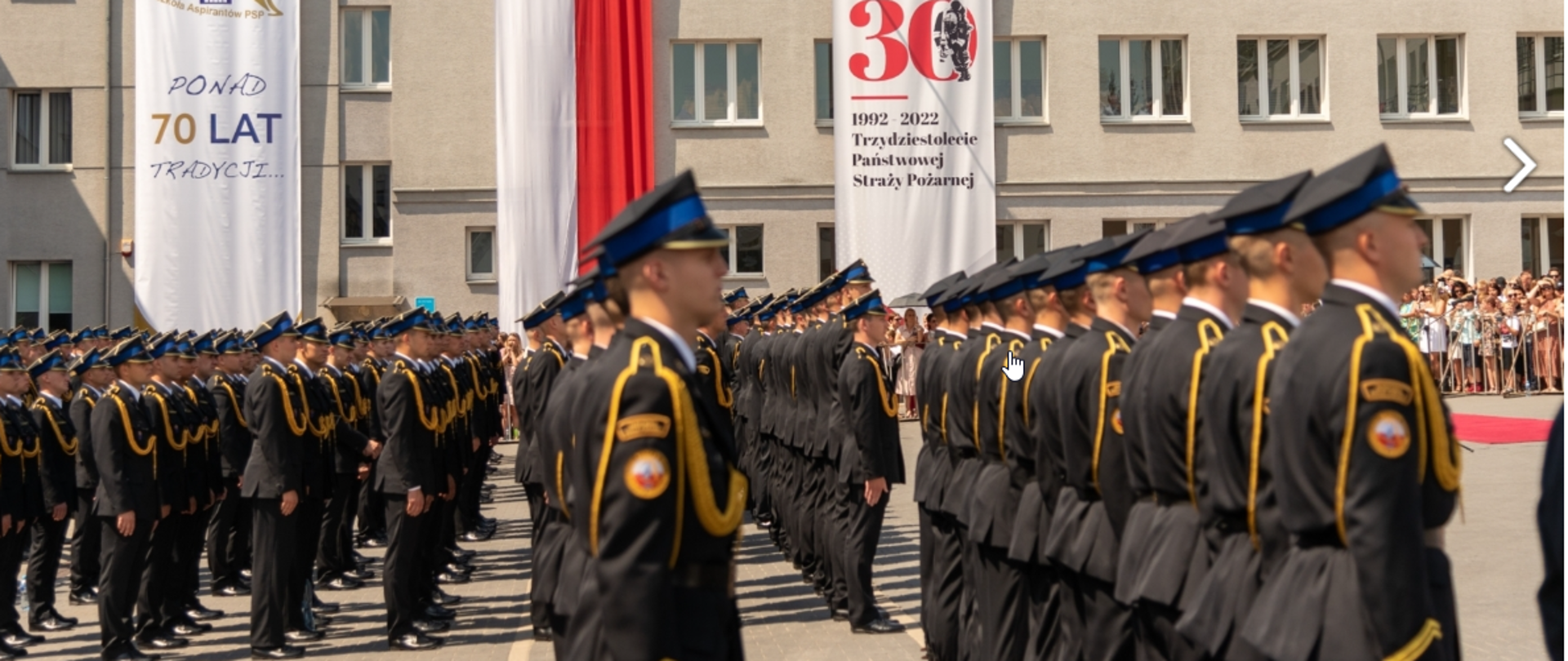 grupa strażaków w ciemnych mundurach, ze sznurem i w rogatywkach stoi w szeregach, w tle flaga biało czerwona rozwiszona na budynku, obok niej dwa banery - jeden z napisem 30 , 1992-2022 trzydziestolecie państwowej straży pożarnej, drugi z napisem poad 70 lat tradycji