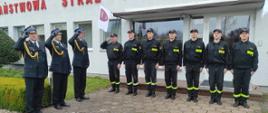 Zdjęcie przedstawia salutujących funkcjonariuszy Państwowej Straży Pożarnej. Fotografia została wykonana przed budynkiem komendy.