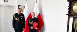 Pomorski komendant wojewódzki Państwowej Straży Pożarnej stoi z funkcjonariuszem po wręczeniu dokumentu. Strażak trzyma czerwoną teczkę. Za nimi stoją flagi państwowe.