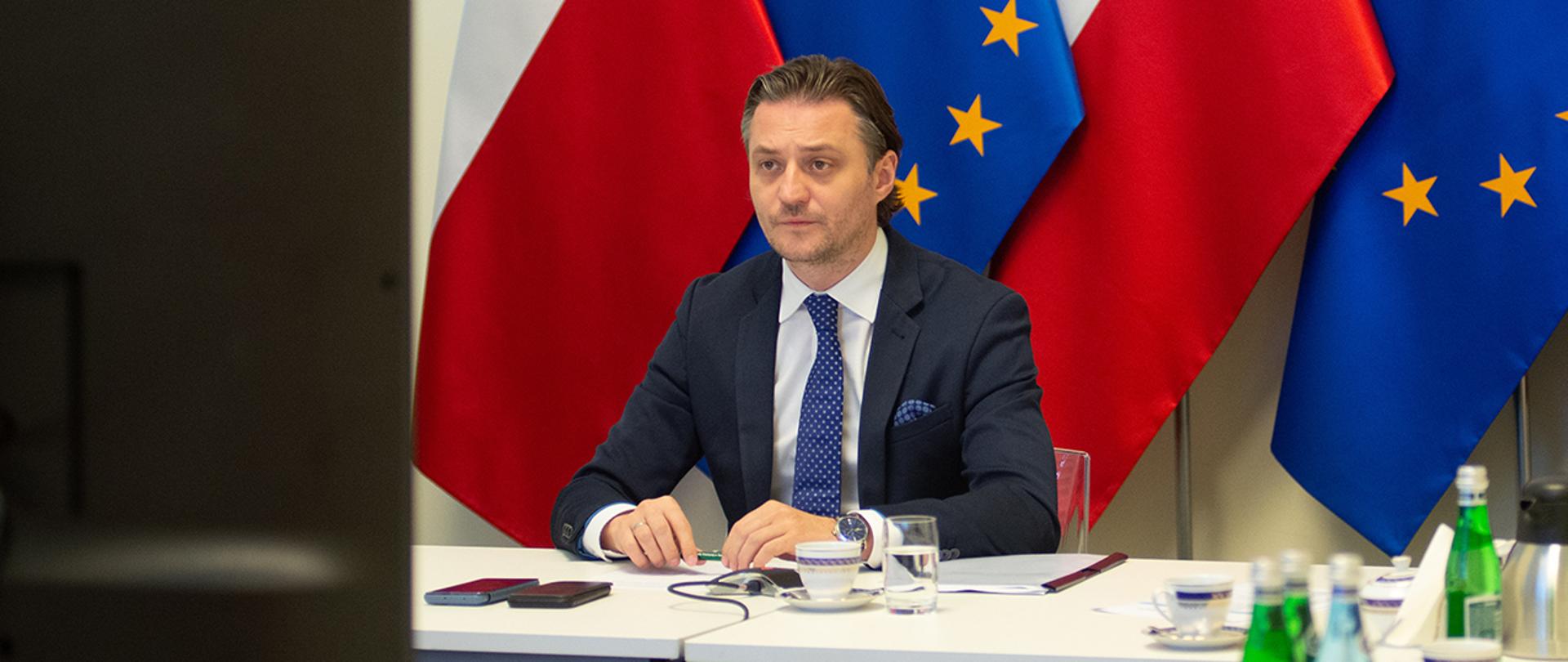 Na zdjęciu widać wiceministra Bartosza Grodeckiego siedzącego za stołem na tle flag Polski i UE.
