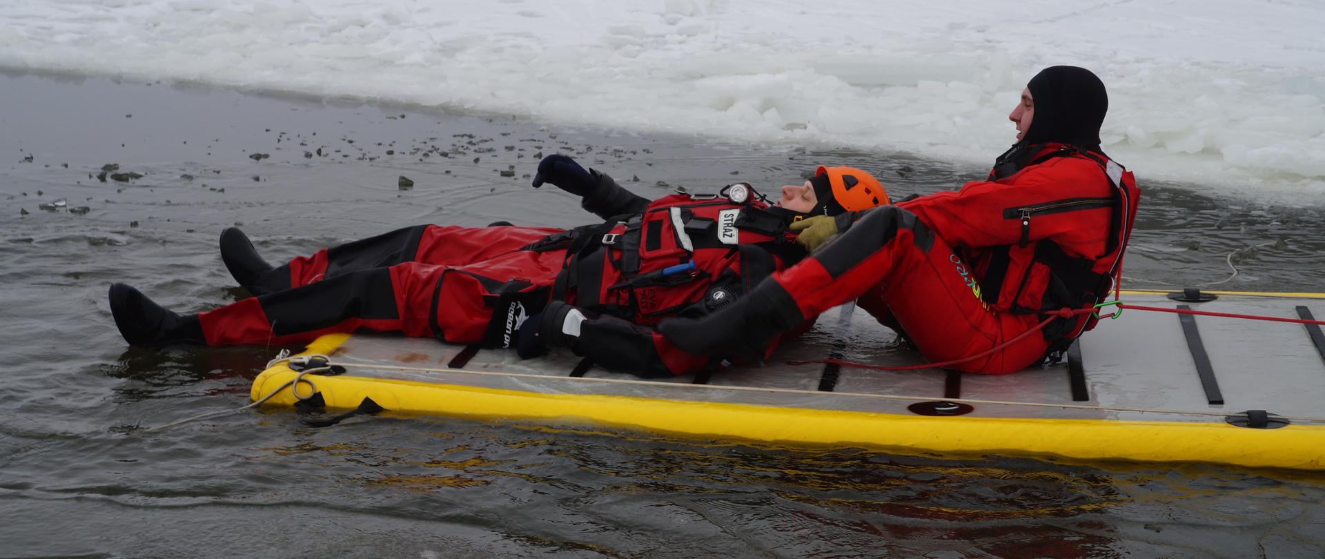 załamany lód, w przerębli znajduje się człowiek, rozwinięty trap ratowniczy na który wciągana przez strażaka jest osoba uwieziona w lodzie