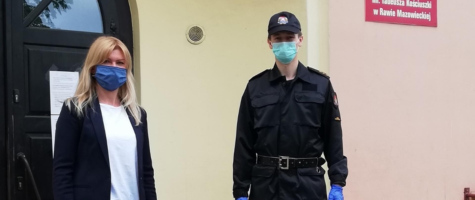 Zdjęcie przedstawia funkcjonariusza PSP w mundurze dowódczo-sztabowy oraz kobietę, blondynkę. Obydwoje są w maseczkach. Między nimi stoją środki do dezynfekcji (5 baniek 5 litrowych). Stoją przed wejściem do budynku Szkoły Podstawowej nr 1 w Rawie Mazowieckiej.