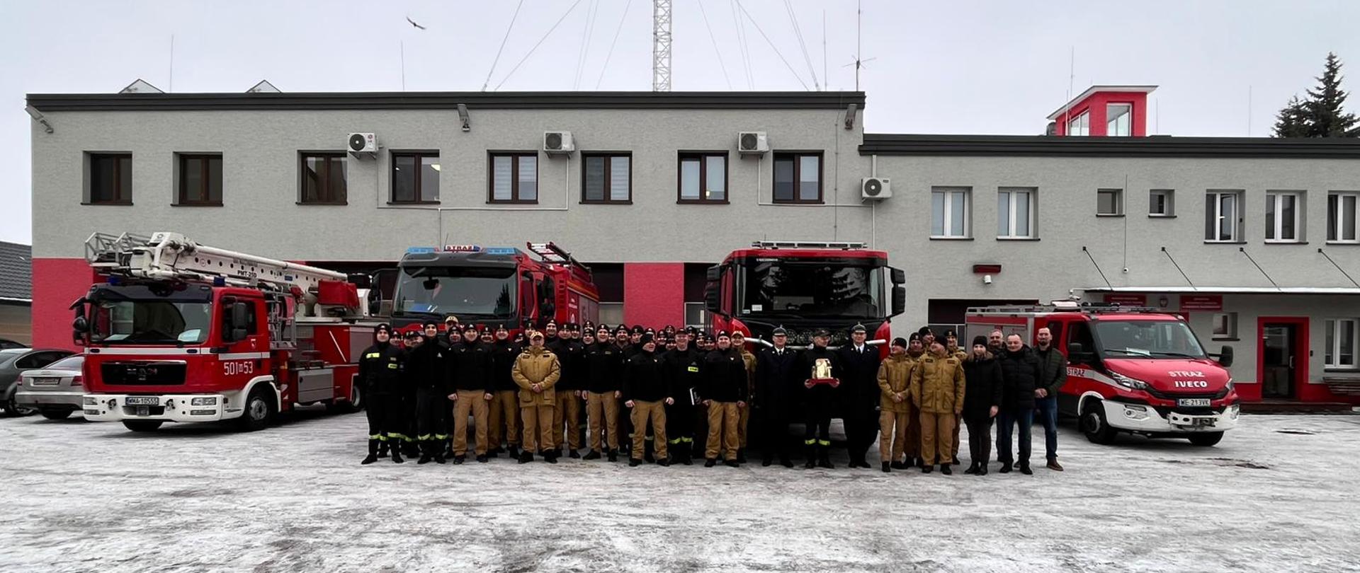 Wspólne zdjęcie funkcjonariuszy i pozostałych osób na zewnątrz komendy, z tyłu stoją wozy strażackie. 