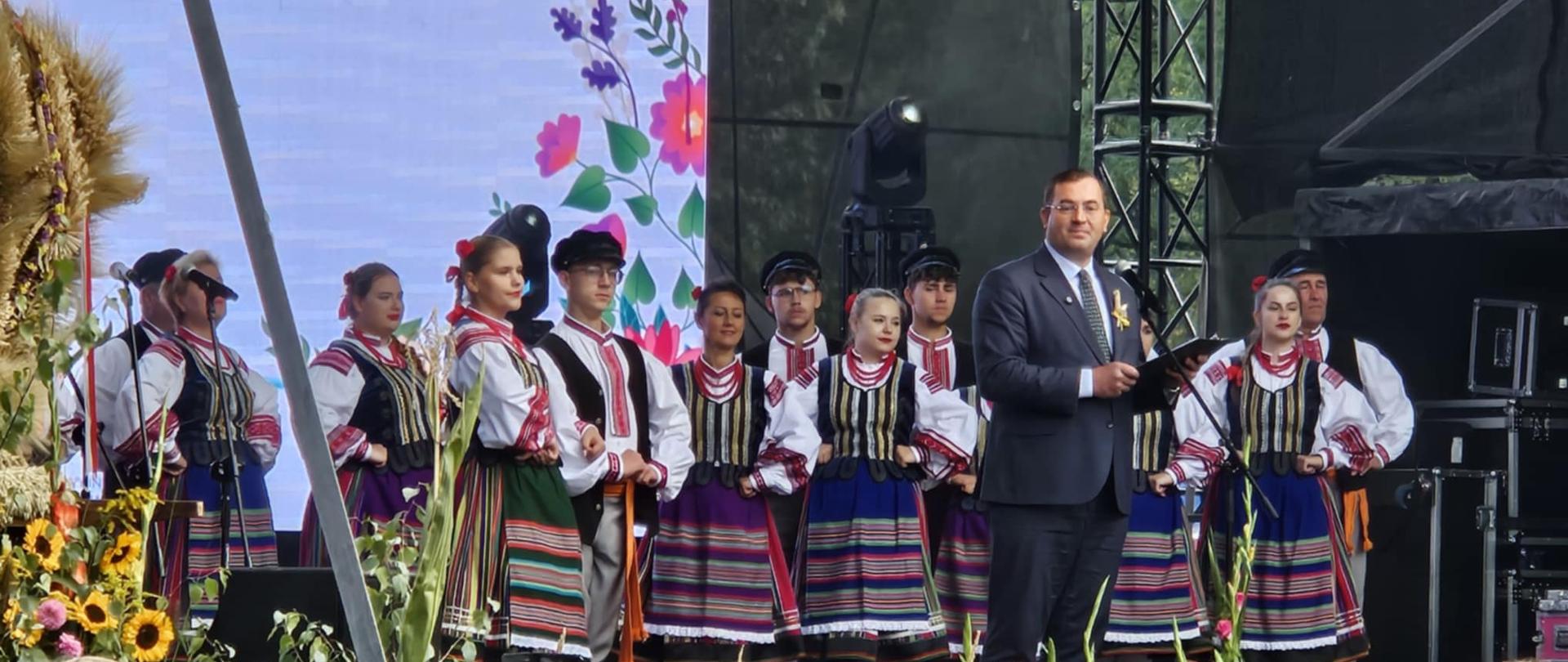 Mężczyzna stojący przy mikrofonie na scenie, w tle członkowie zespołu ludowego.