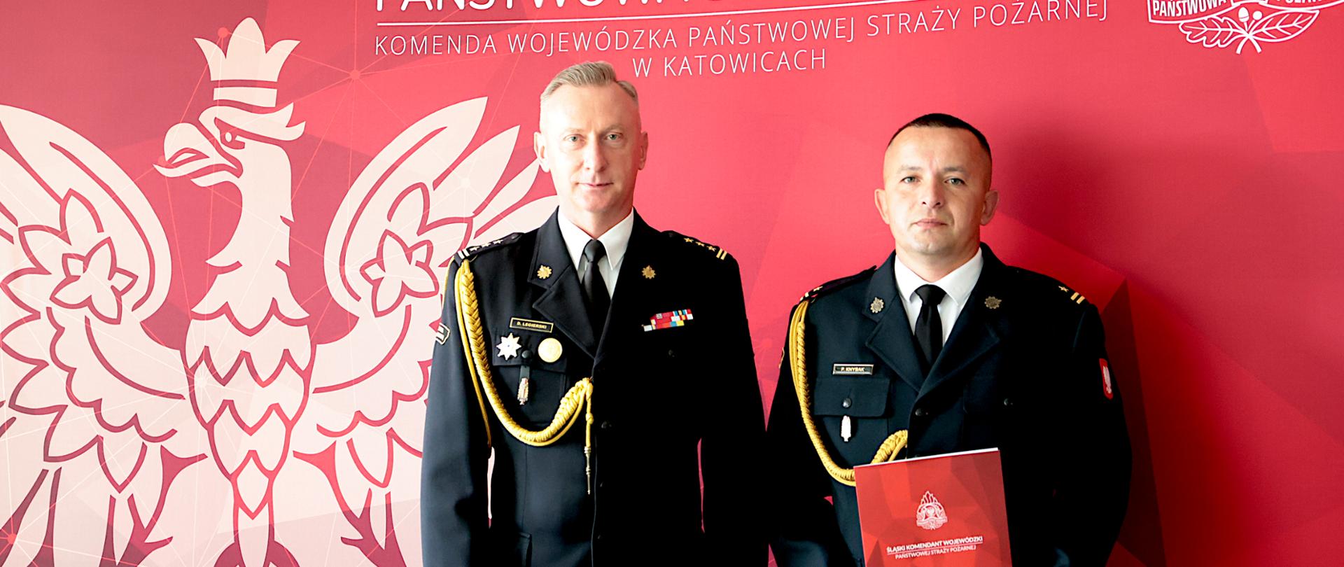 Dwaj funkcjonariusze Państwowej Straży Pożarnej w ciemnych galowych mundurach stoją obok siebie i patrzą w stronę aparatu. Ich mundury zdobią dystynkcje, odznaczenia oraz złote sznury galowe, a mężczyzna po prawej trzyma przed sobą czerwoną teczkę z emblematem straży. W tle znajduje się czerwona ścianka z dużym białym wizerunkiem orła, godłem formacji oraz napisem „Państwowa Straż Pożarna – Komenda Wojewódzka Państwowej Straży Pożarnej w Katowicach”.