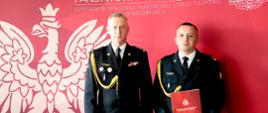 Dwaj funkcjonariusze Państwowej Straży Pożarnej w ciemnych galowych mundurach stoją obok siebie i patrzą w stronę aparatu. Ich mundury zdobią dystynkcje, odznaczenia oraz złote sznury galowe, a mężczyzna po prawej trzyma przed sobą czerwoną teczkę z emblematem straży. W tle znajduje się czerwona ścianka z dużym białym wizerunkiem orła, godłem formacji oraz napisem „Państwowa Straż Pożarna – Komenda Wojewódzka Państwowej Straży Pożarnej w Katowicach”.