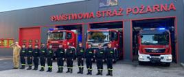 W bramie garażu budynku Państwowej Straży Pożarnej stoją trzy samochody ratowniczo - gaśnicze z włączonymi sygnałami świetlnymi, przed samochodami w szeregu stoją w ubraniach koszarowych funkcjonariusze oddając hołd zmarłemu druhowi
