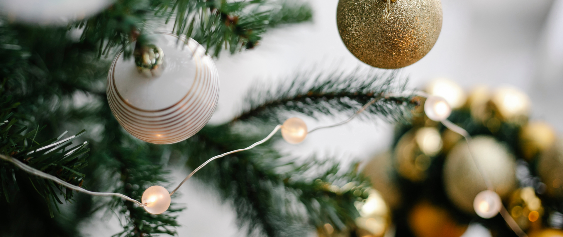 Zbliżenie na gałązki choinki ozdobione białą bombką w złote paski, złotą brokatową bombką oraz zapalonym sznurem lampek o ciepłym świetle. W tle widoczne rozmyte świąteczne dekoracje.