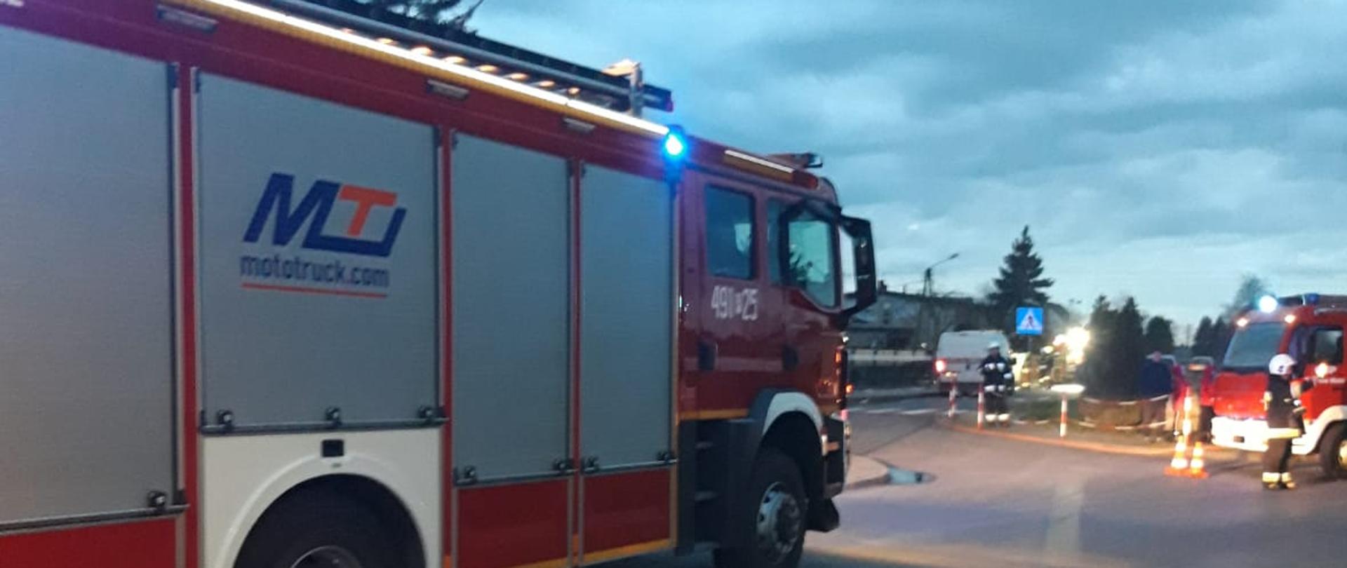 Zdjęcie przedstawia wozy straży pożarnej oraz strażaków stojących na drodze podczas zabezpieczania miejsca prowadzonych działań.