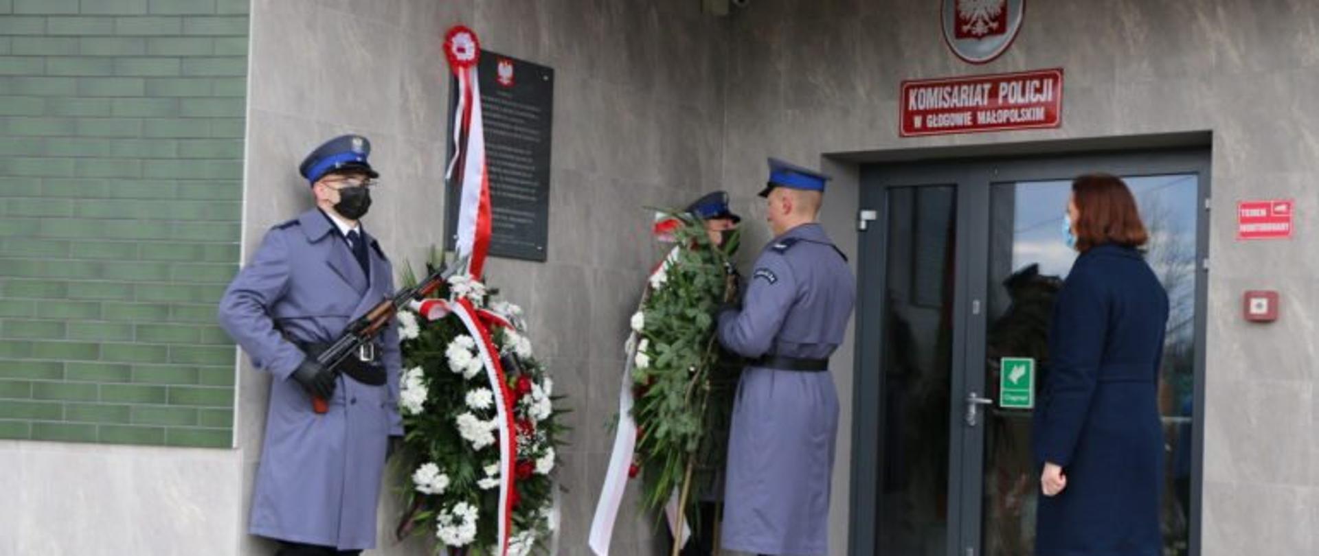 Wojewoda podkarpacki Ewa Leniart stoi przed tablicą upamiętniającą funkcjonariuszy Policji zamordowanych przez NKWD. Przed nią stoi policjant trzymający, wieniec. Tablica znajduje się na ścianie budynku komisariatu Policji w Głogowie Małopolskim