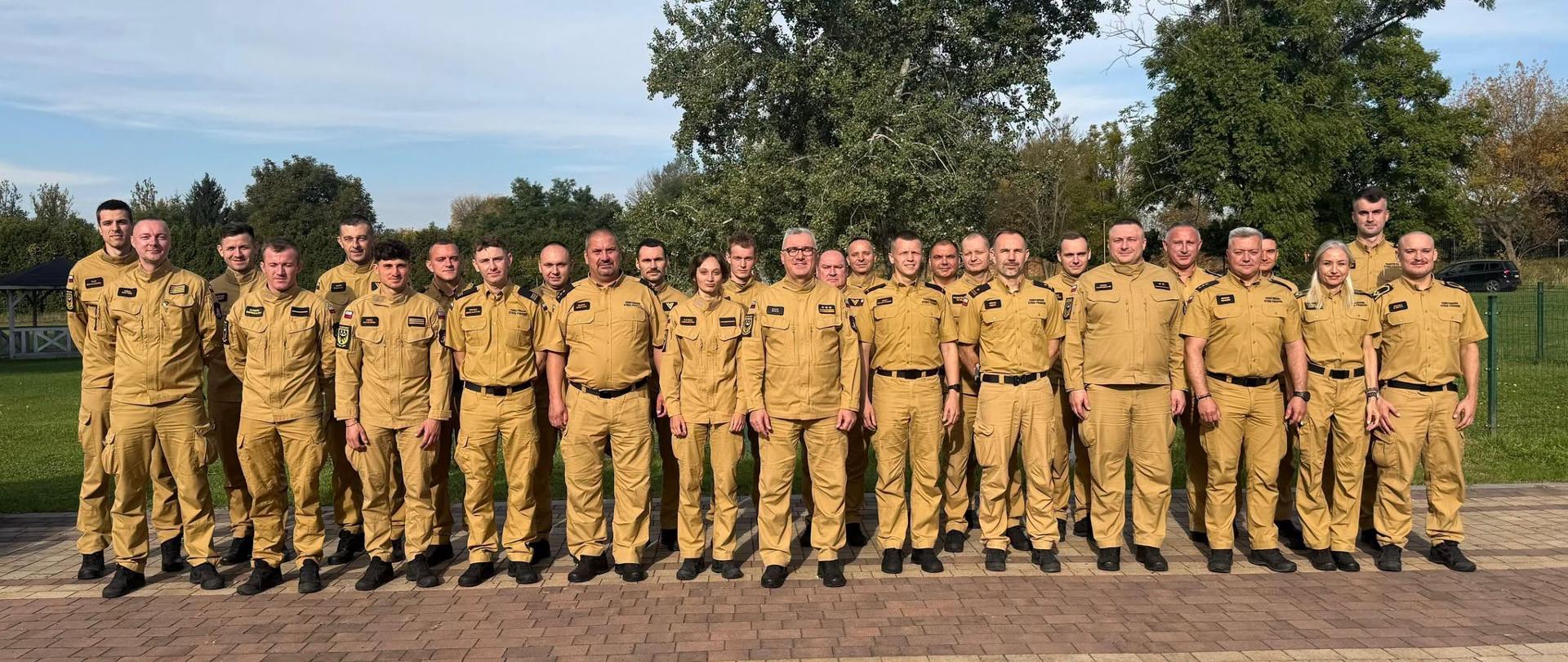 Grupa strażaków i pracowników Państwowej Straży Pożarnej pozuje do pamiątkowego zdjęcia na zewnątrz, stojąc w kilku rzędach na tle zieleni i drzew. Wszyscy uczestnicy ubrani są w jednolite jasnobrązowe (piaskowe) mundury służbowe z pasami i czarnym obuwiem. W kadrze widoczni są zarówno mężczyźni, jak i kobiety w różnym wieku, stojący na wyłożonym kostką brukową placu.