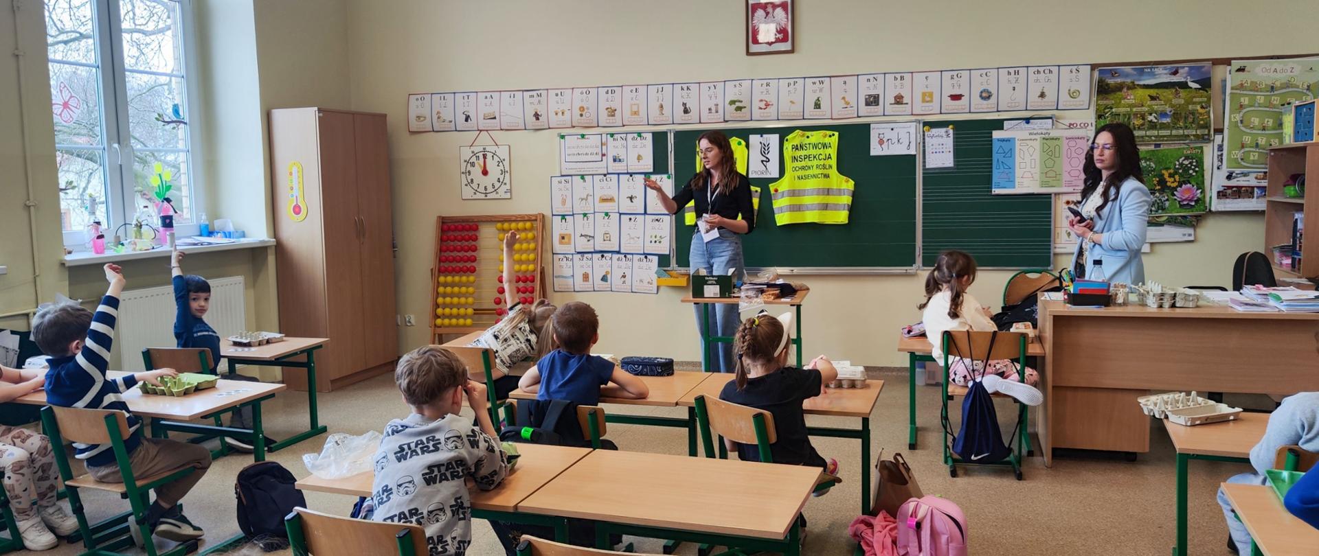 W klasie lekcyjnej przed uczniami siedzącymi w ławkach stoi pełniąca obowiązki kierownika Oddziału w Lubaniu pani Marta Wyszyńska prowadząca zajęcia. Troje dzieci podnosi rękę do góry zgłaszając się.