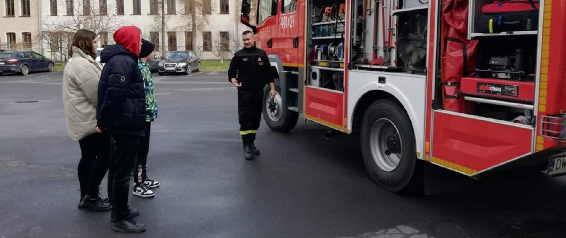 Obraz przedstawia strażaka prezentującego samochód z sprzętem pożarniczym dla grupy dzieci