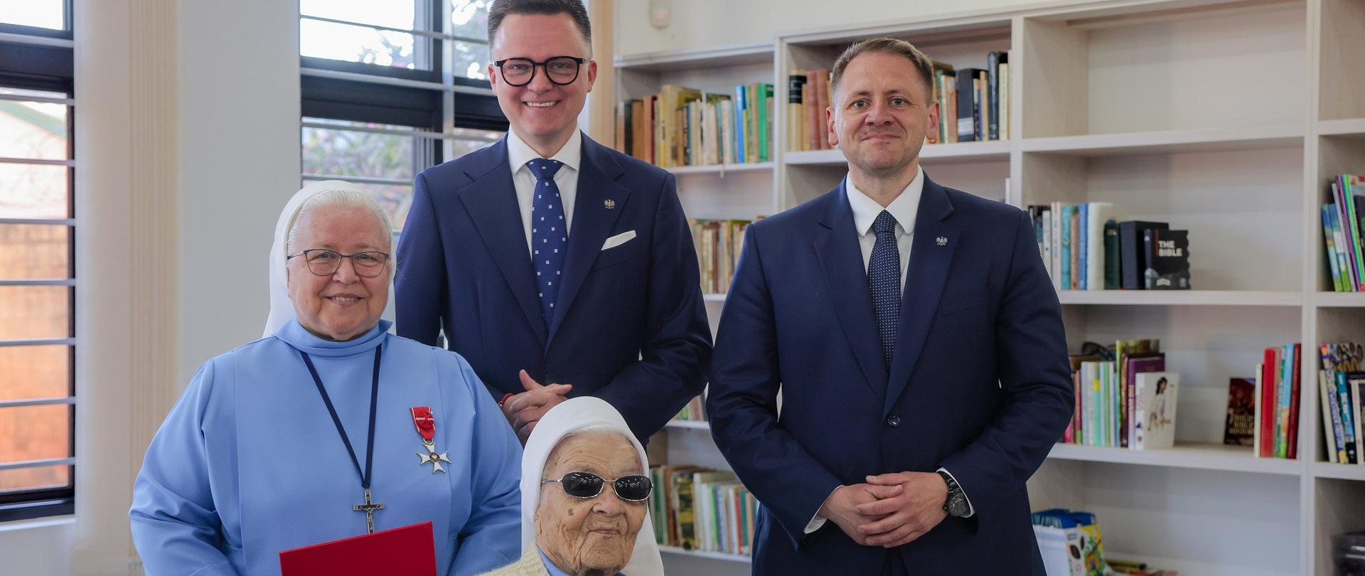 Grupa czterech osób, na pierwszym planie dwie siostry zakonne, z odznaczeniami państwowymi (Krzyż Komandorski Orderu Odrodzenia Polski i Krzyż Oficerski Orderu Odrodzenia Polski), jedna z sióstr siedzi w fotelu inwalidzkim, na drugim planie stoi dwóch mężczyzn: Wicemarszałek Sejmu Szymon Hołownia oraz Podsekretarza Stanu Mateusz Kotecki; w tle regały z książkami i dwa zakratowane okna