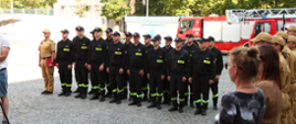 Strażacy Państwowej Straży Pożarnej w mundurach kolory czarnego oraz piaskowego wraz z osobami cywilnymi stoją na baczność za nimi stoi samochód specjalny drabina.