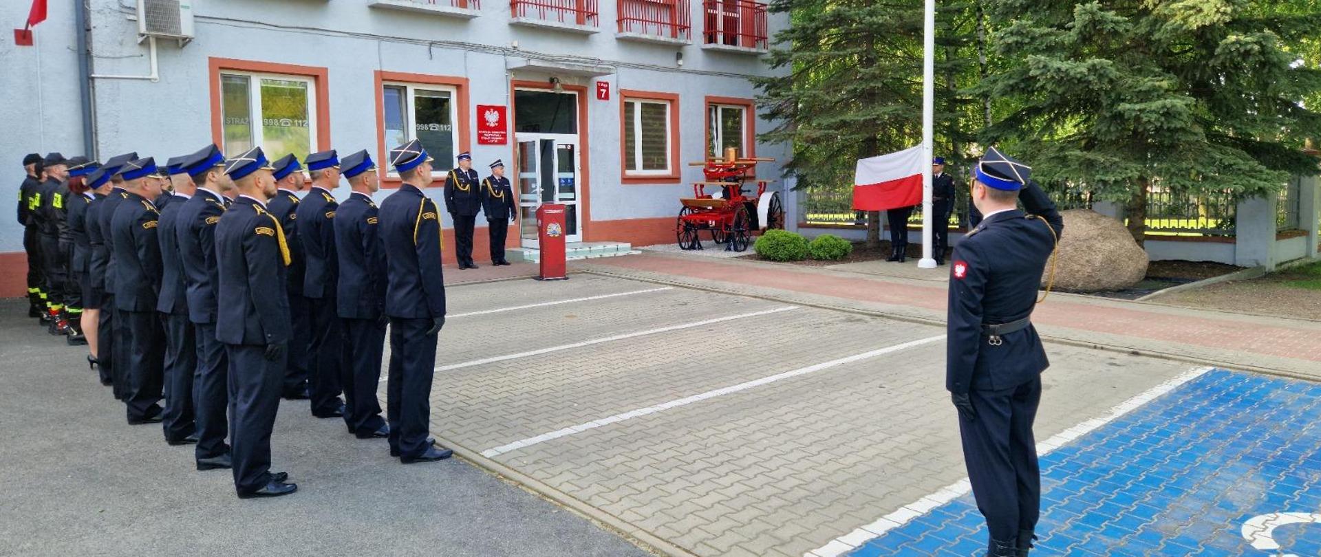 strażacy na uroczystej zmianie służby przed budynkiem komendy powiatowej, przy maszcie flagowym stoi poczet flagowy