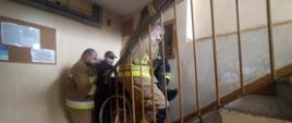 Zdjęcie przedstawia trzech strażaków z jednostki OSP Wleń oraz strażnika miejskiego, którzy pomagają znieść starszą osobę na wózku inwalidzkim po schodach w budynku. 