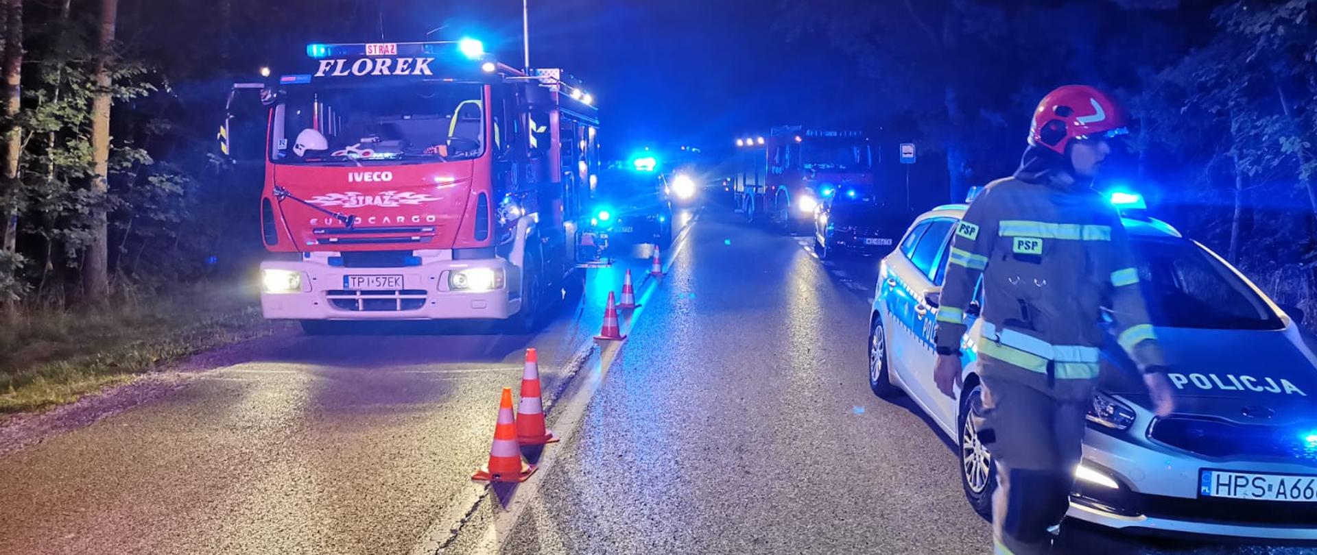 Zdjęcie przedstawia samochody pożarnicze oraz radiowóz obecne na miejscu zdarzenia. Obok pojazdów widać strażaka PSP w umundurowaniu bojowym.