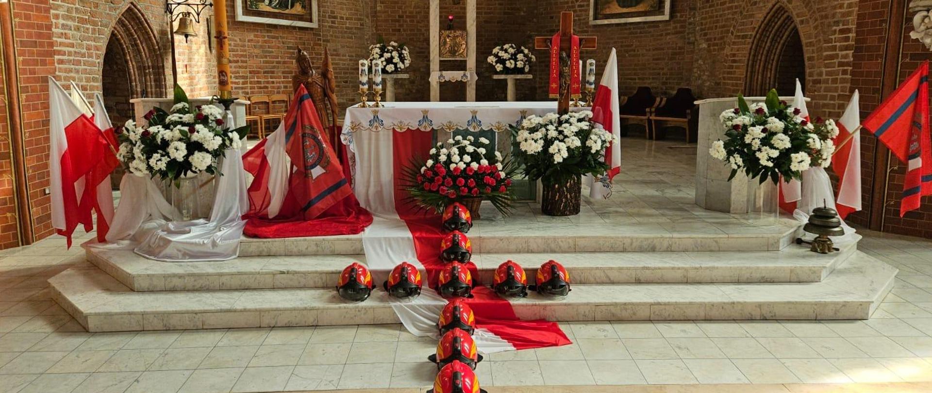 Ołtarz w kościele przystrojony we flagi biło czerwone, Na schodach leżą czerwone hełmy strażackie na kształt krzyża