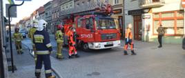 Pożar kamienicy na rawickim rynku. Ulica grunwaldzka. Na środku ulicy stoi specjalny wóz strażacki z podnośnikiem hydraulicznym. Wokół niego znajduje się pięciu strażaków oraz 3-osobowa załoga zespołu ratownictwa medycznego. W tle budynki i samochody. 