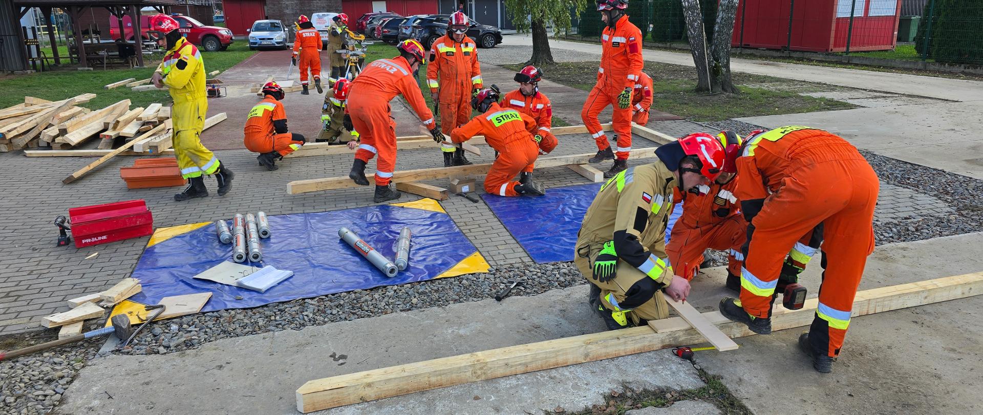 Ratownicy, głównie w pomarańczowych i jeden w żółto-zielonym kombinezonie, pracują na utwardzonym placu. Część z nich klęczy i układa drewniane elementy konstrukcyjne, inni zajmują się sprzętem leżącym na niebieskich plandekach. W tle widać budynki i zaparkowane samochody.
