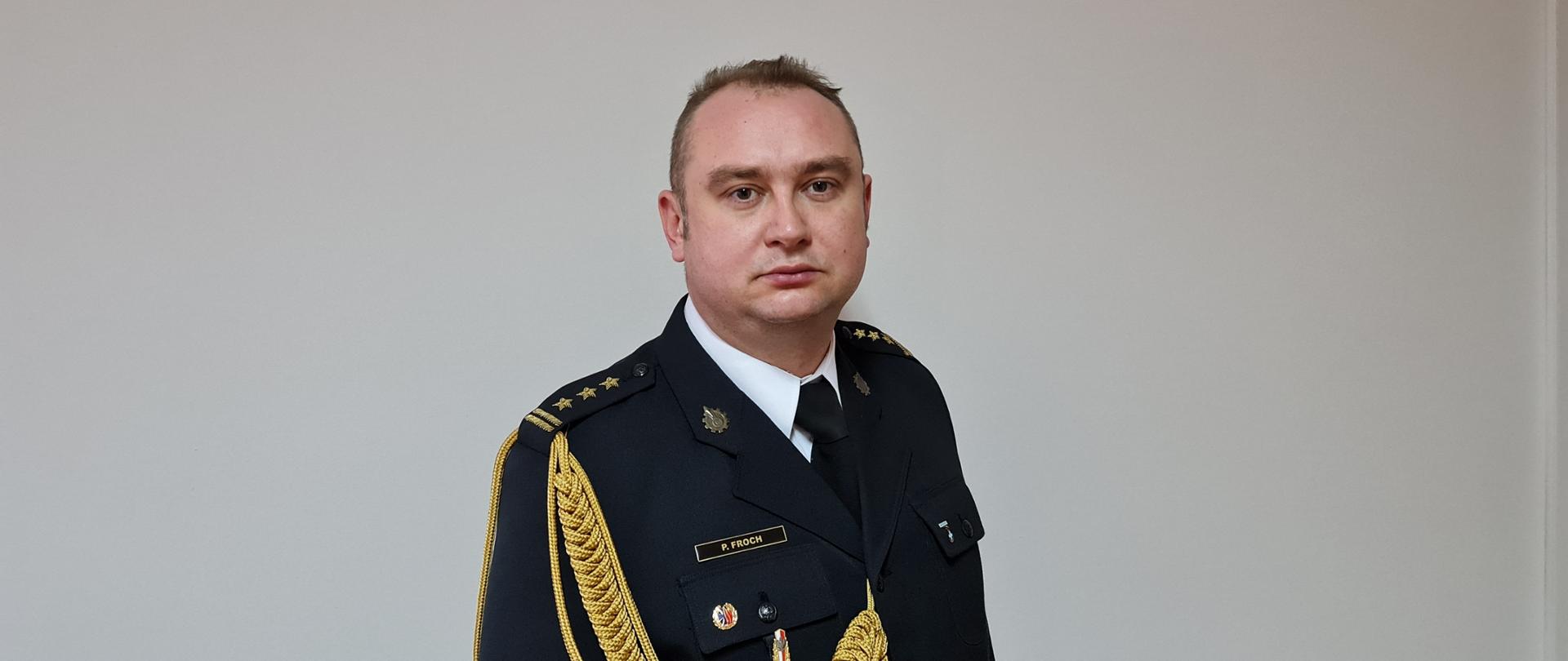 Na zdjęciu widać mężczyznę w mundurze galowym, stojącego na tle jasnej ściany. Ma on krótkie, jasne włosy i patrzy prosto przed siebie. Ubrany jest w ciemny mundur, ozdobiony złotymi pagonami z trzema gwiazdkami oraz złotym, plecionym sznurem przewieszonym przez lewe ramię. Na lewej piersi munduru widoczne są przypinki. Mężczyzna ma poważny wyraz twarzy.