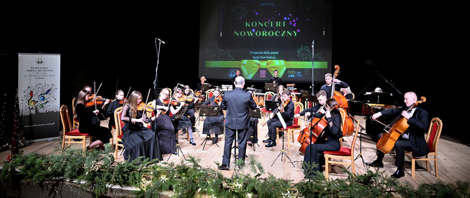 Zdjęcie. Na estradzie Sali Widowiskowej Nyskiego Domu Kultury występuje orkiestra. Z przodu estrada przyozdobiona gałązkami iglaków. w tle baner promujący Koncert Noworoczny. Z lewej strony w tle stoi baner PSM Nysa. Zespół prowadzi dyrygent.